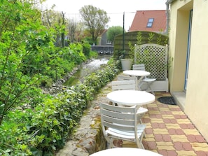Un espace patio aménagé avec des meubles d’extérieur blancs, un petit ruisseau et une végétation luxuriante.
