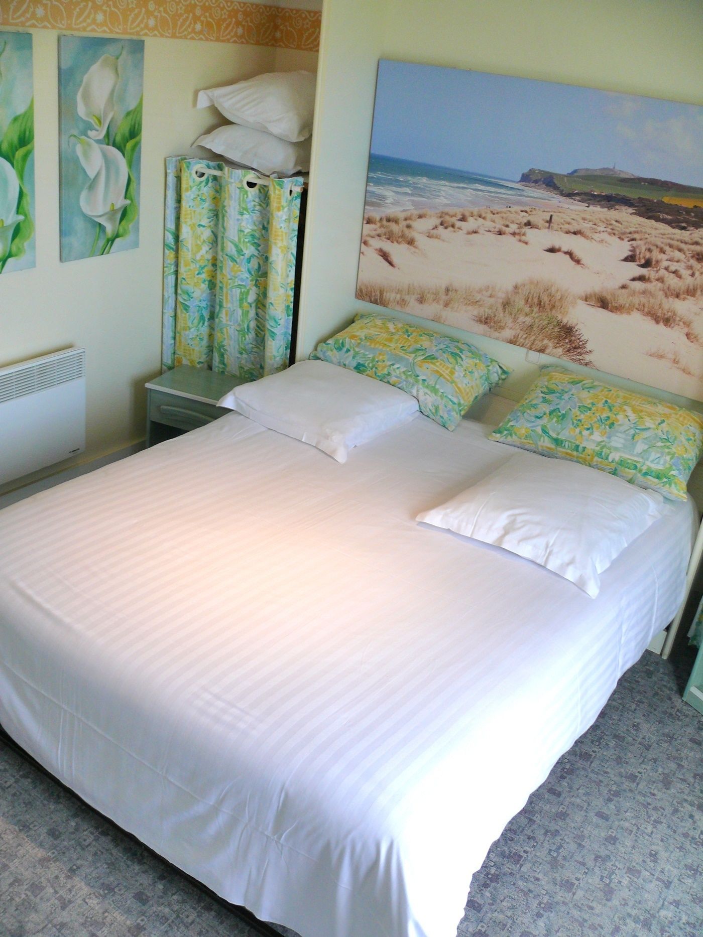 Uma cama bem arrumada com lençóis brancos, uma pequena mesa de cabeceira com uma luminária, e um quadro com uma paisagem de praia na parede.