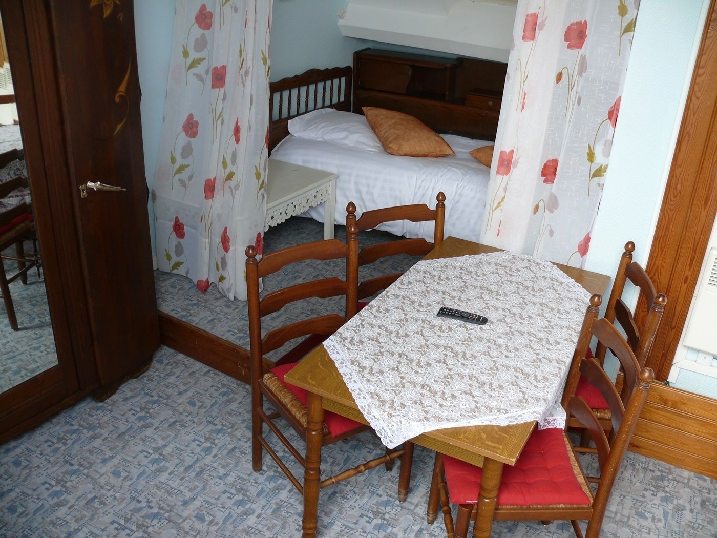 A bedroom with a bed, a chair, a table with a remote control, and floral curtains.