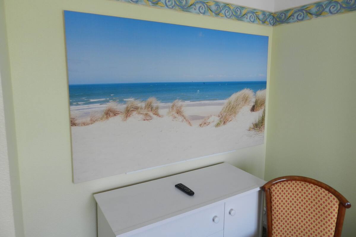 Una habitación con un cuadro grande de playa en la pared, un tocador blanco y una silla de madera.