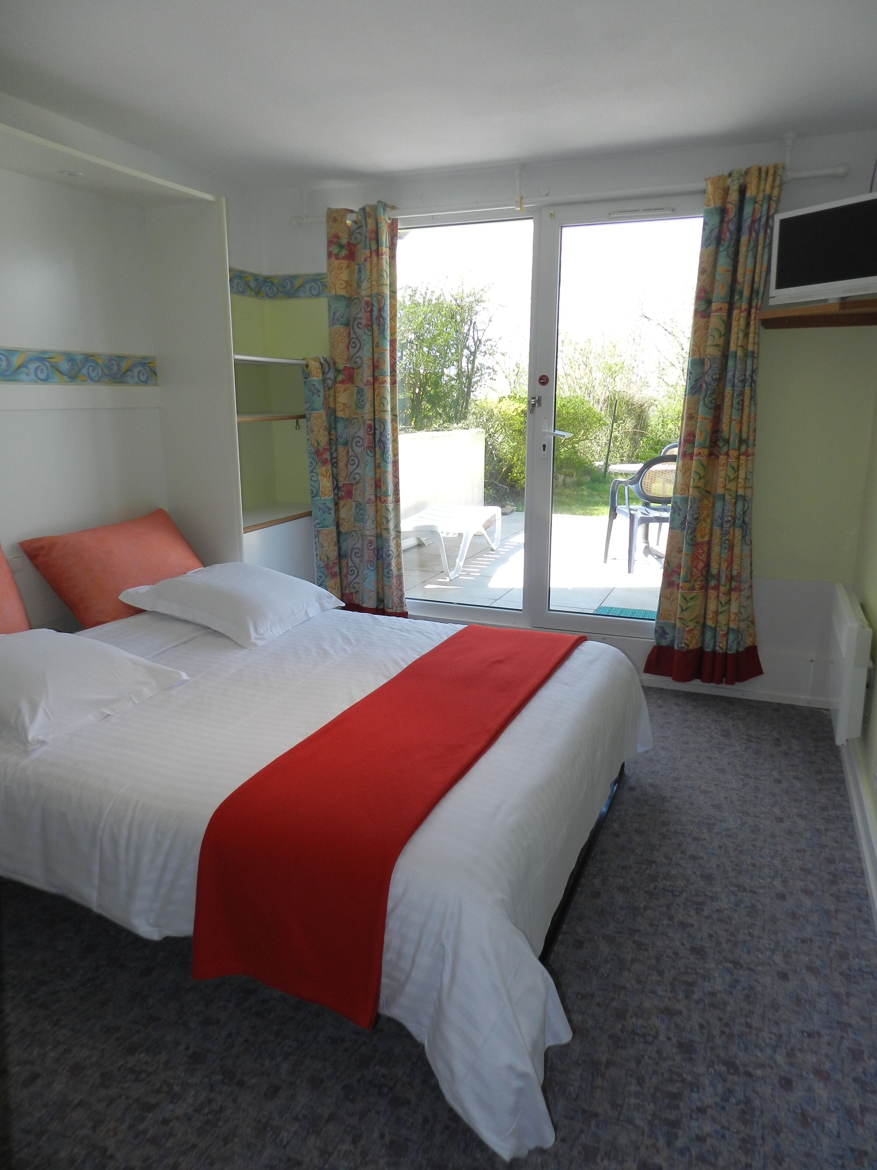 Ein Hotelzimmer mit einem Bett, einer roten Decke, einem Fenster mit Vorhängen und Blick ins Freie.