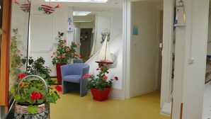 A hallway with a blue chair, potted plants with red flowers, a small sailboat decoration, and a white wall with a clock.