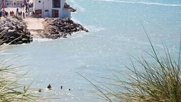 Un bâtiment situé sur la côte, avec une rive rocheuse, une vue dégagée sur la mer et quelques personnes dans l’eau.