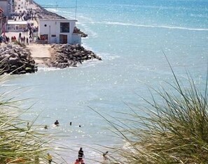Un edificio costero con un litoral rocoso, vista despejada al mar y algunas personas en el agua.