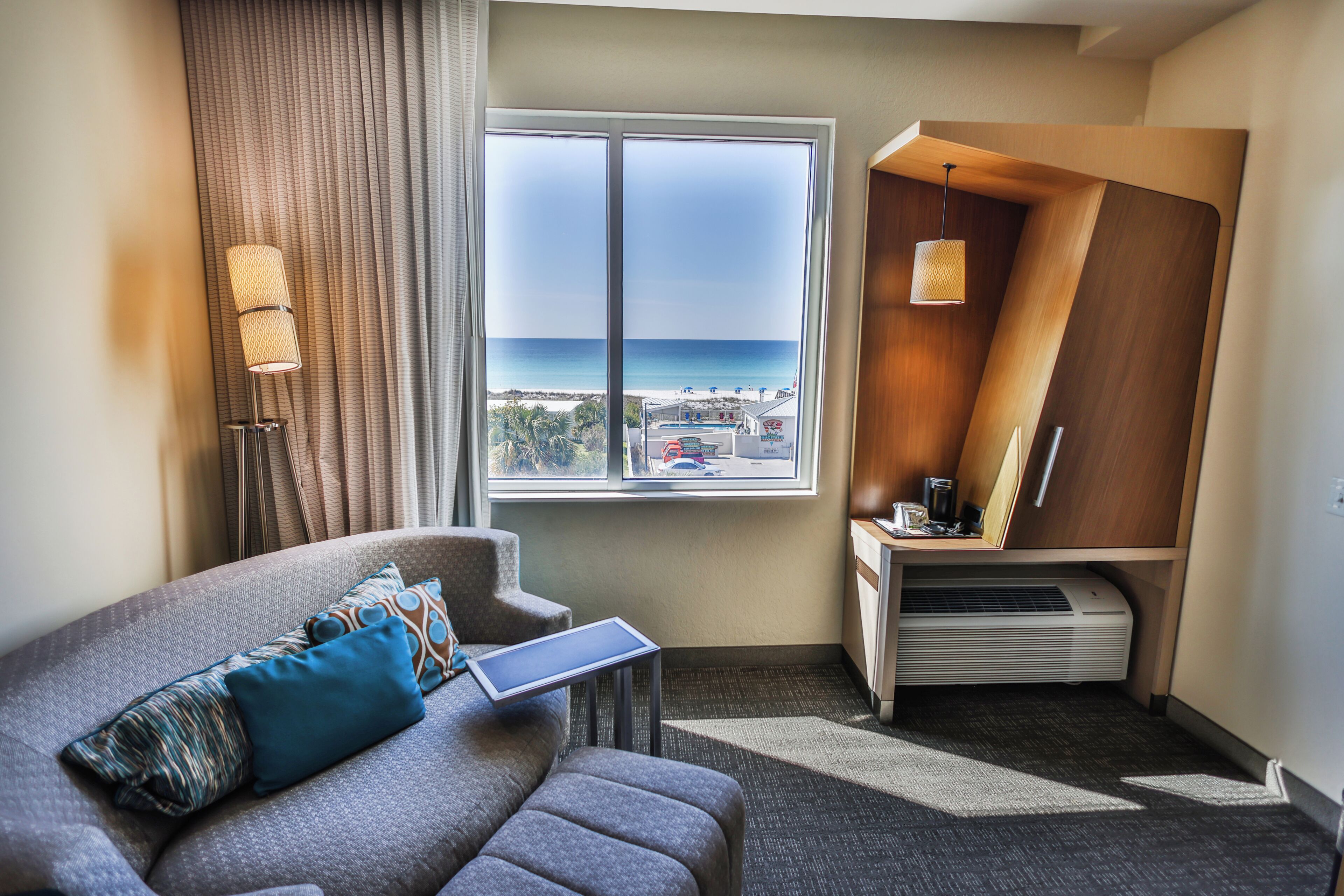 Habitación de hotel con vista a la playa, un sofá con un ordenador portátil, un escritorio con una lámpara y un equipo de aire acondicionado integrado.