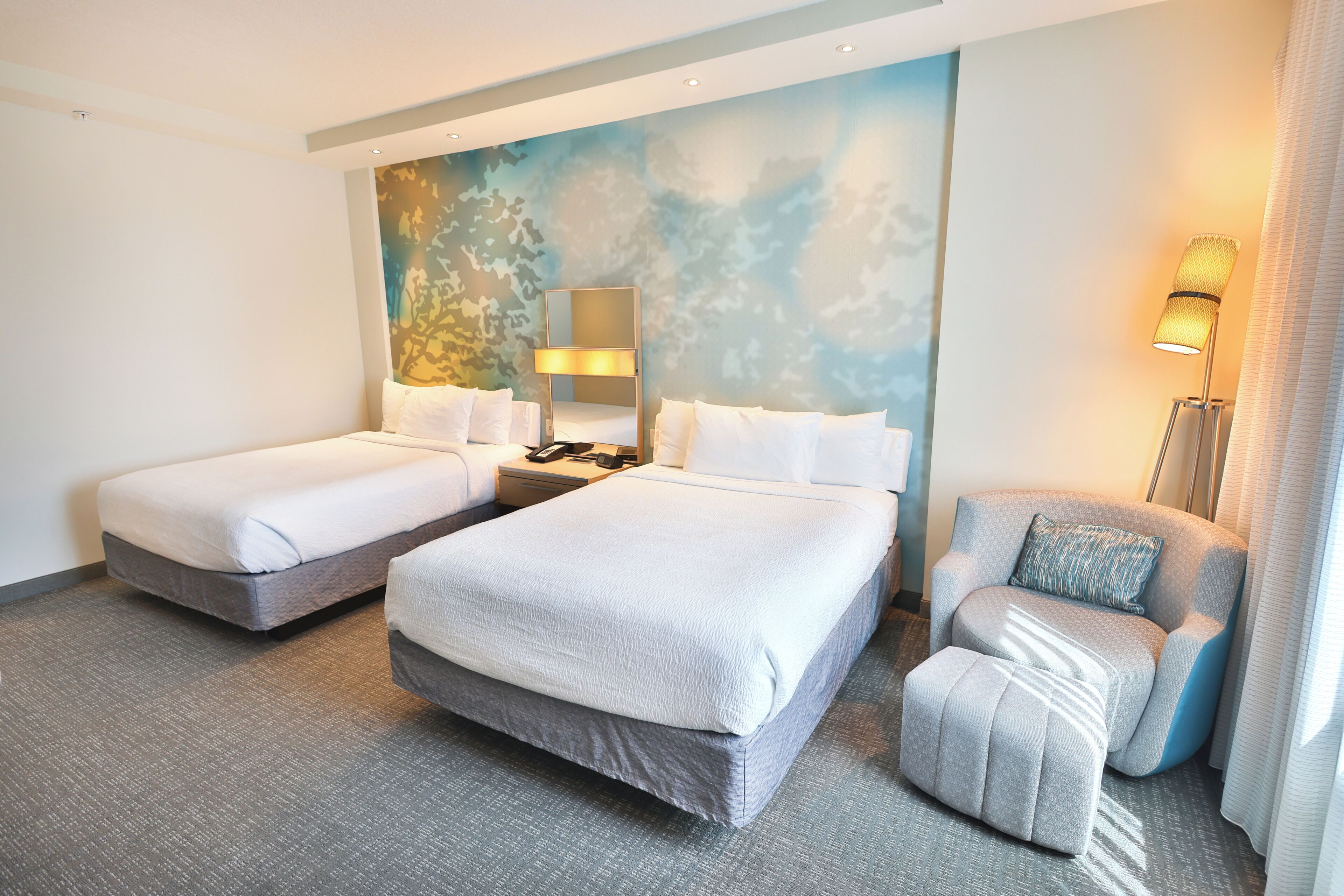 A hotel room with two beds, a chair, a nightstand, and a wall mural.