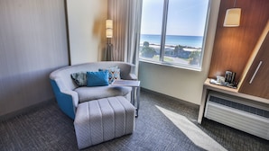 A hotel room with a view of the ocean, a grey sofa with blue cushions, a small table, a lamp, and a heater.