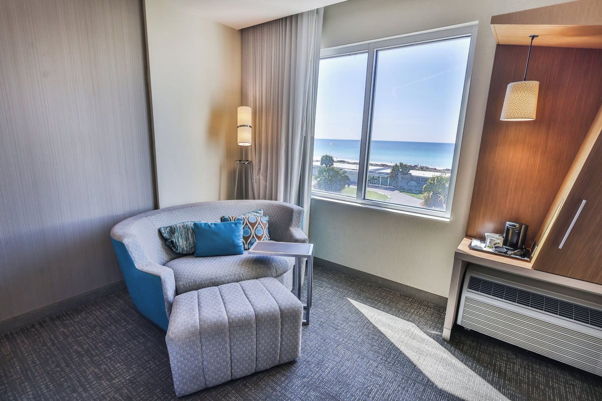 Een hotelkamer met uitzicht op de oceaan, een grijze bank met blauwe kussens, een klein tafeltje, een lamp en een verwarming.