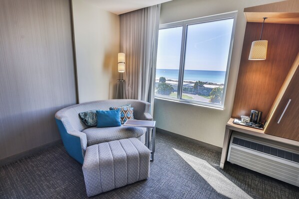 Ett hotellrum med utsikt över havet, en grå soffa med blå kuddar, ett litet bord, en lampa och en värmare.