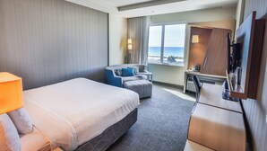 A hotel room with a bed, a desk, a chair, a TV, and a window with a view of the ocean.