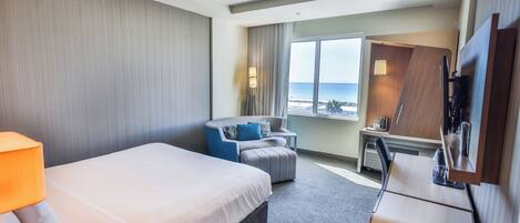 A hotel room with a bed, a desk, a chair, a TV, and a window with a view of the ocean.