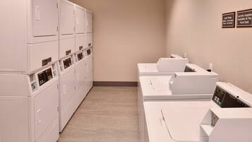 Une buanderie équipée de plusieurs machines à laver et de plusieurs sèche-linge, avec un mur de couleur beige et un panneau indiquant que le nécessaire pour faire la lessive est disponible à la réception.