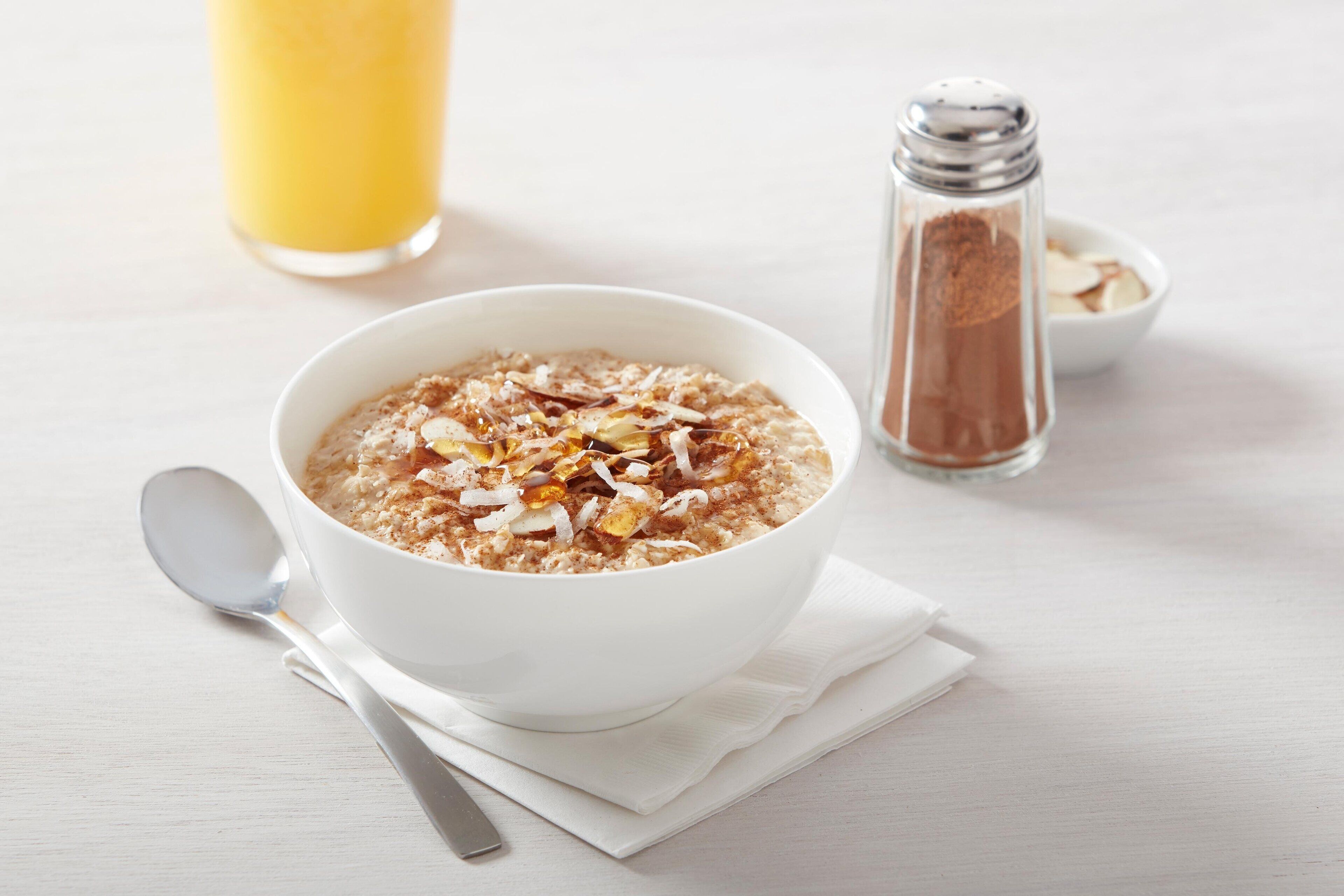 A bowl of oatmeal with almonds and coconut flakes, a salt shaker, and a glass of orange juice on a white table.