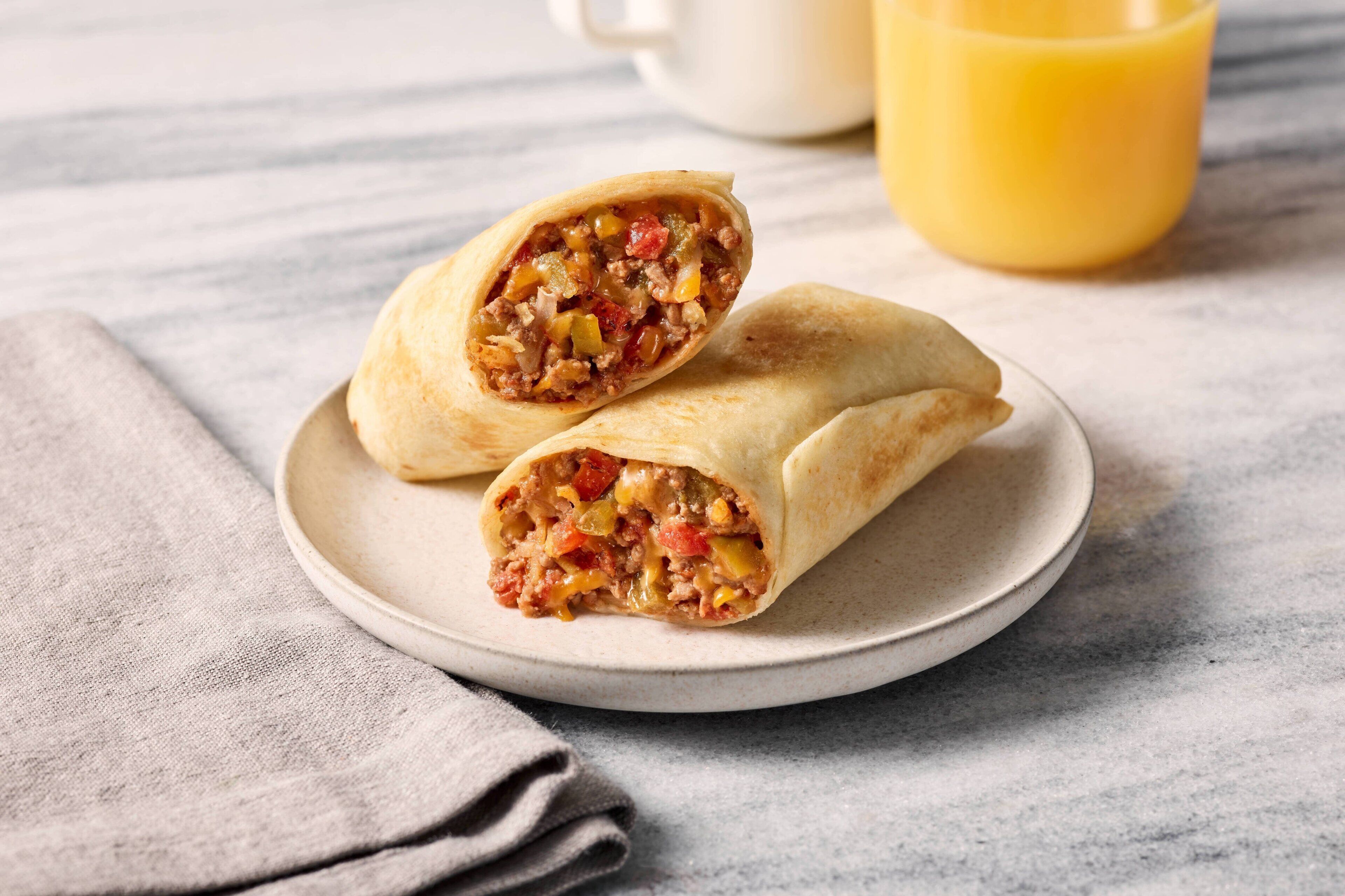 A burrito on a plate with a cup of orange juice in the background.