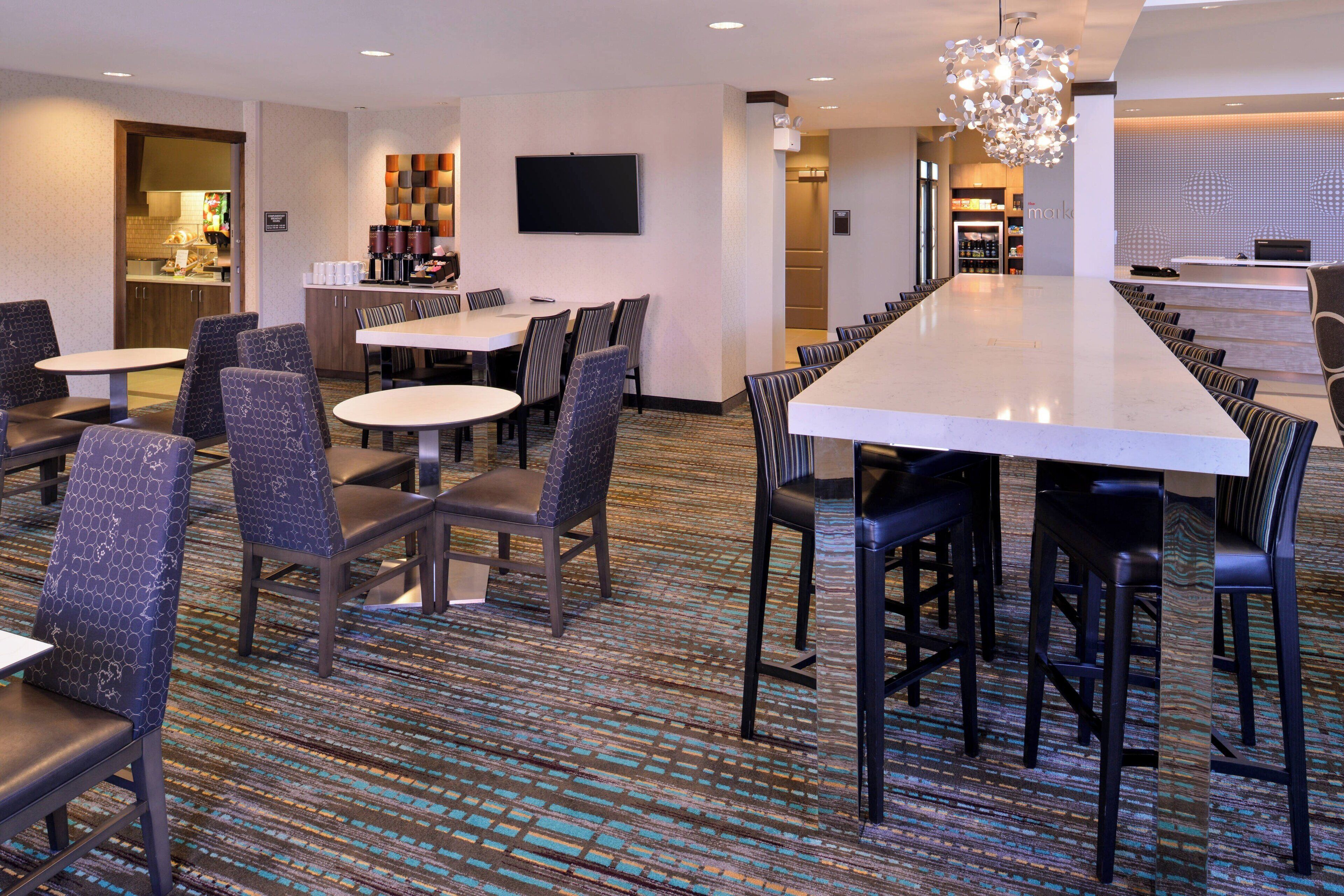 A hotel conference room with a long table, multiple chairs, a television, and a kitchen area in the background.