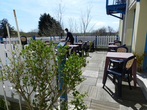 An outdoor patio with wooden tables and chairs, a white fence, and a person walking in the background. - Hôtel Le Vivier WISSANT - Centre Village (Wissant)