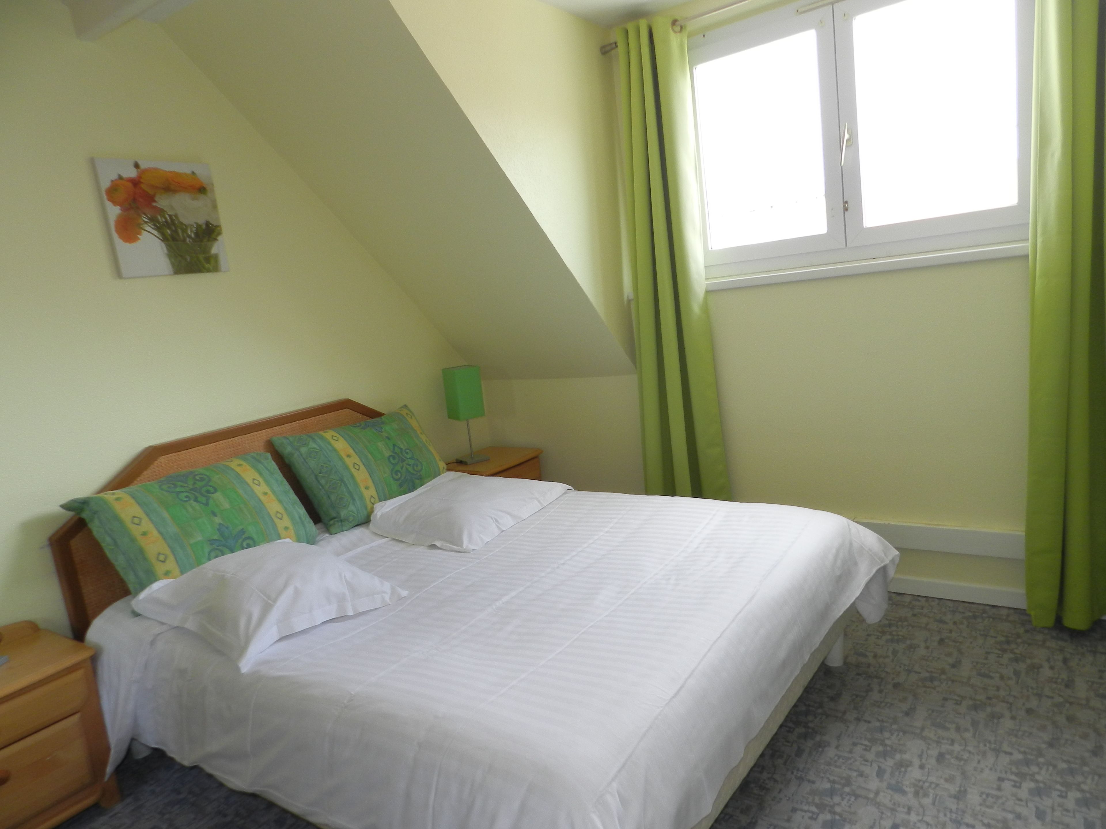 A neatly made bed with white linens in a room with a sloped ceiling and a window with green curtains.