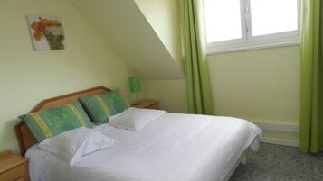 A neatly made bed with white linens in a room with a sloped ceiling and a window with green curtains.