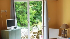 Quarto com cama, televisão em móvel, luminária e vista para o verde por uma porta aberta.