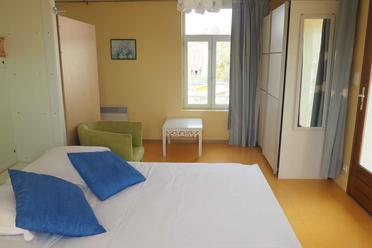 A bedroom with a bed, a green chair, a small table, a window with curtains, and a mirror.