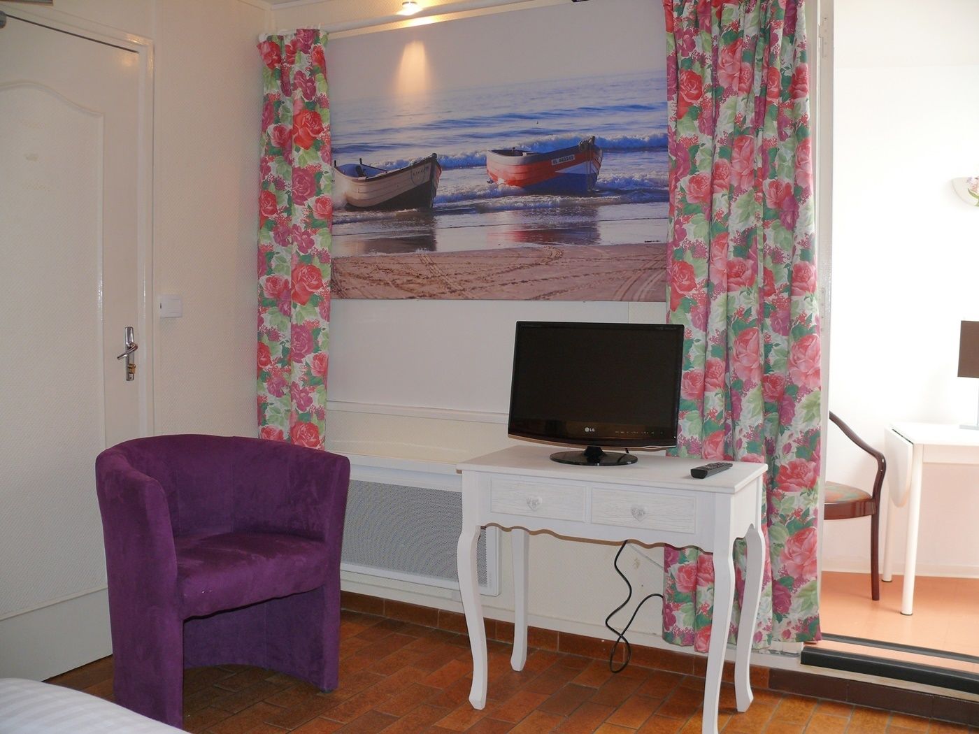 Una habitación con una cama, un sillón morado, un escritorio blanco con un televisor y un cuadro de barcos en la pared.
