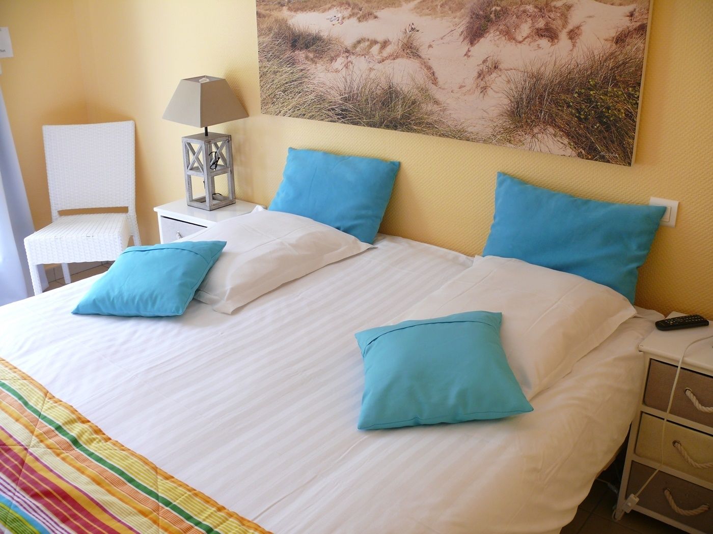 Una cama bien hecha con ropa de cama blanca y azul, una lámpara en una mesita de noche y un cuadro con una playa en la pared.