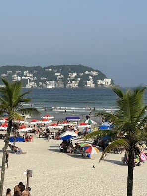 A beach with umbrellas, palm trees, and a view of a coastal town. - Villa Di Verona (Guarujá)