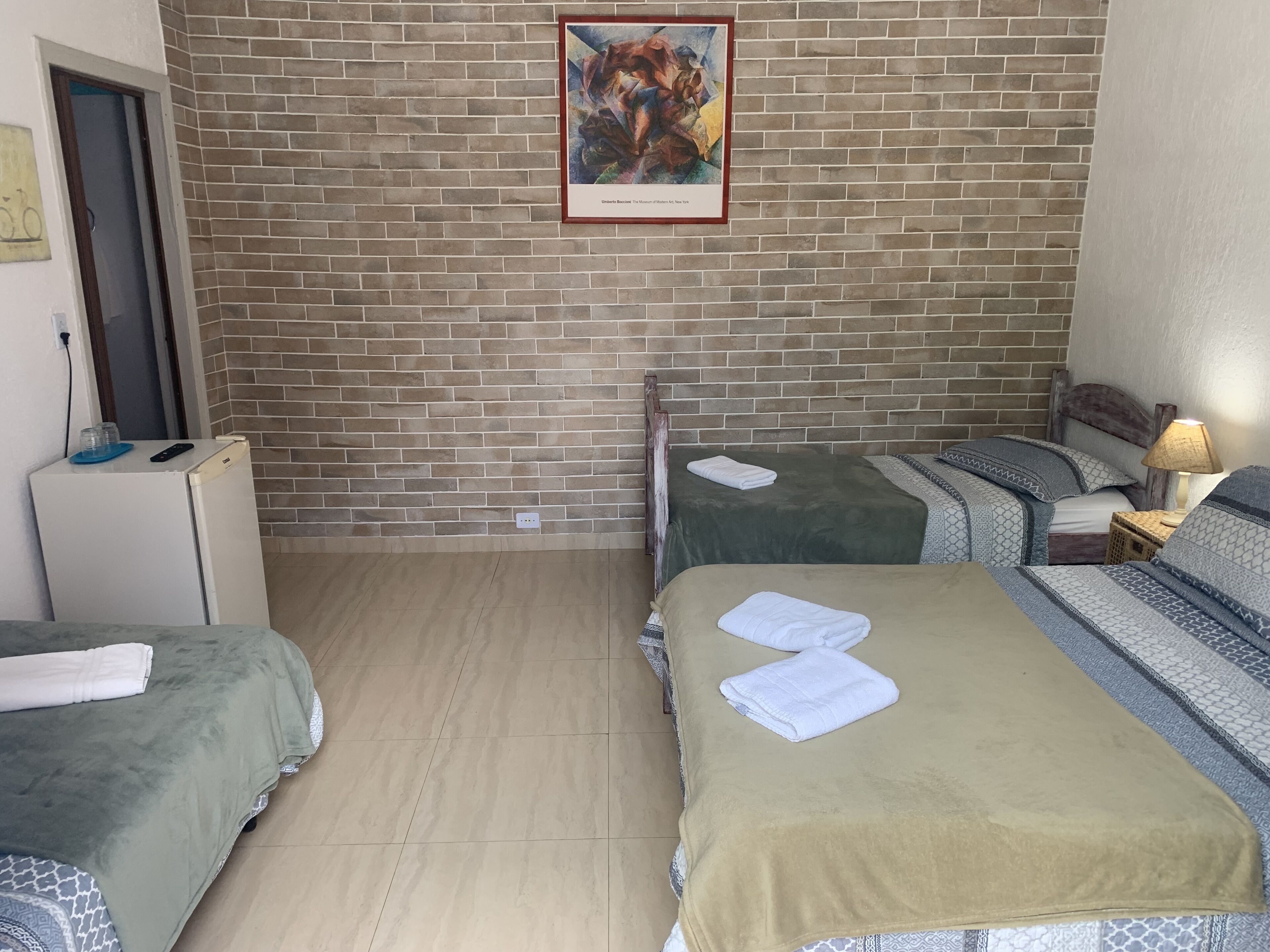 A hotel room with two beds, a brick wall, a painting, a small refrigerator, and a bedside lamp.