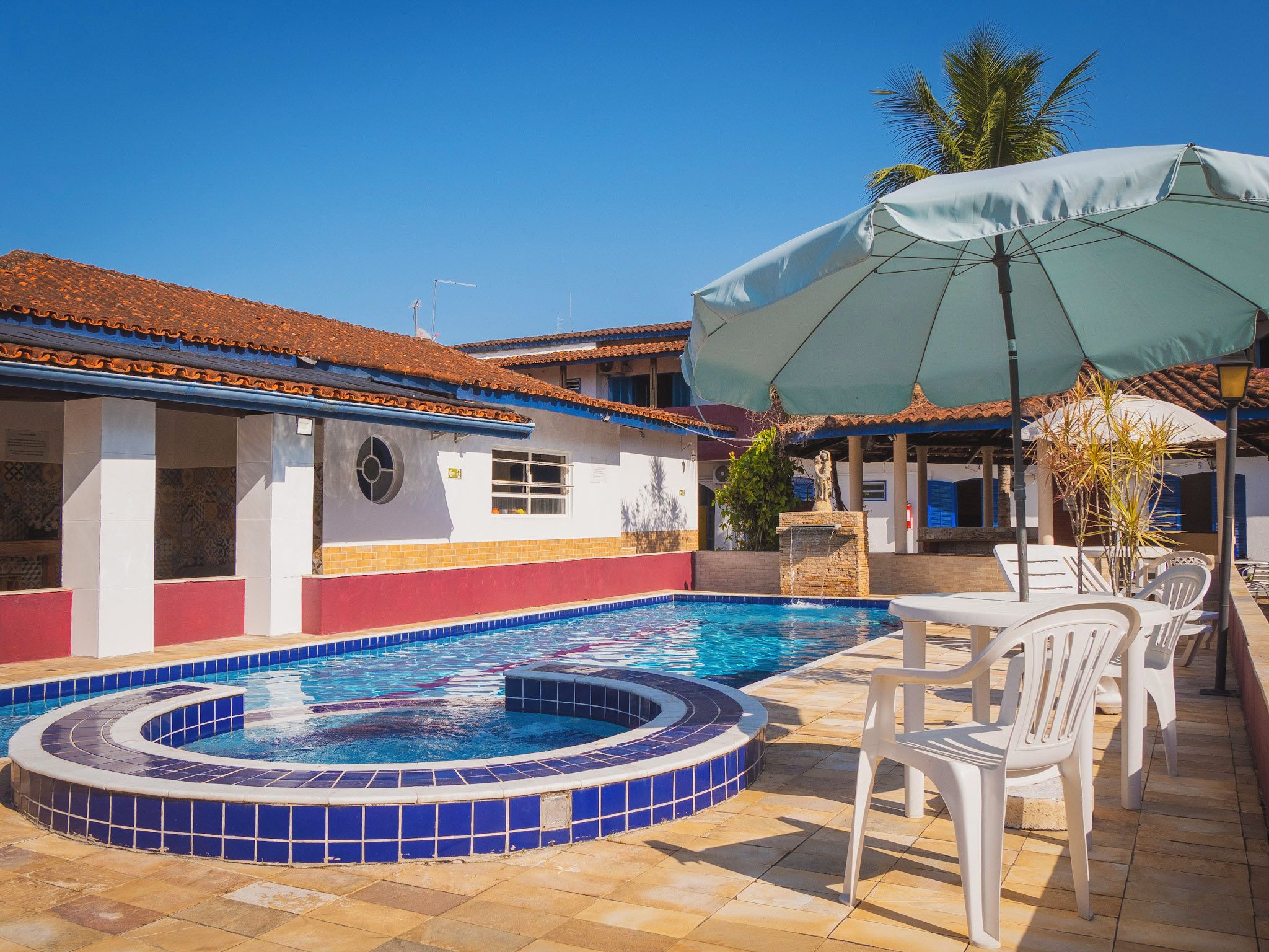 A swimming pool with a tiled edge, surrounded by a patio area with white plastic chairs and a large umbrella.