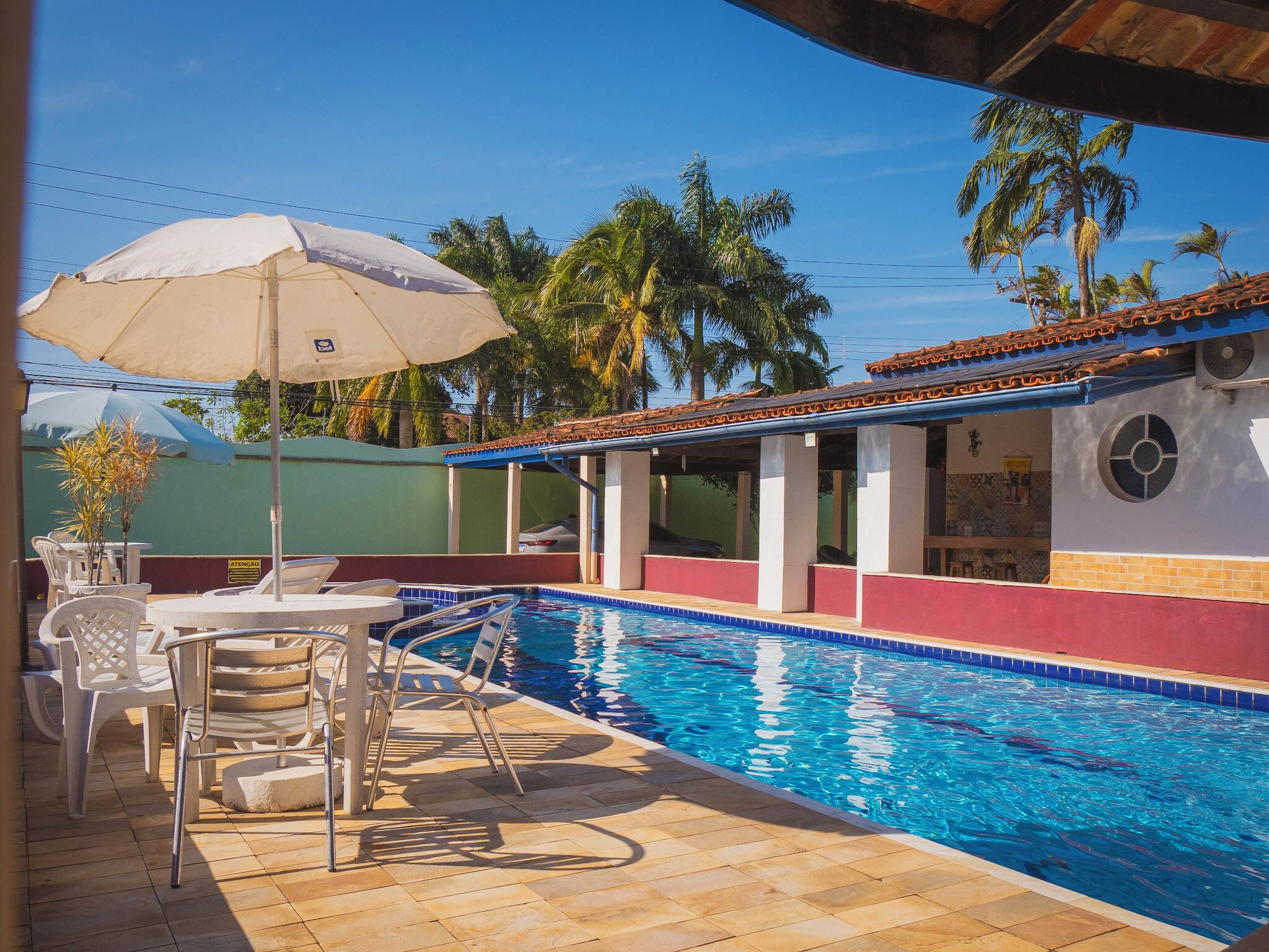 Una piscina con borde de azulejos, rodeada de sillas de descanso y sombrillas, con palmeras al fondo.