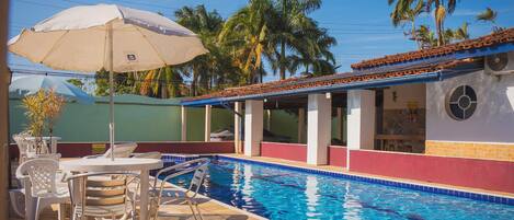 Una piscina con borde de azulejos, rodeada de sillas de descanso y sombrillas, con palmeras al fondo.