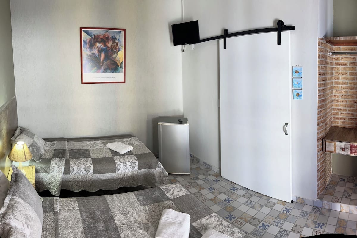 A hotel room with a bed, a small desk, a TV mounted on the wall, a door, and a brick-patterned wall.