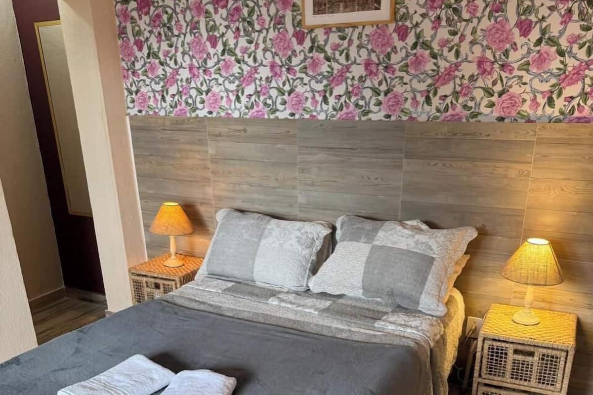 A neatly made bed with a grey bedspread and pillows, two bedside lamps, a wicker bench, and a floral wallpaper pattern.