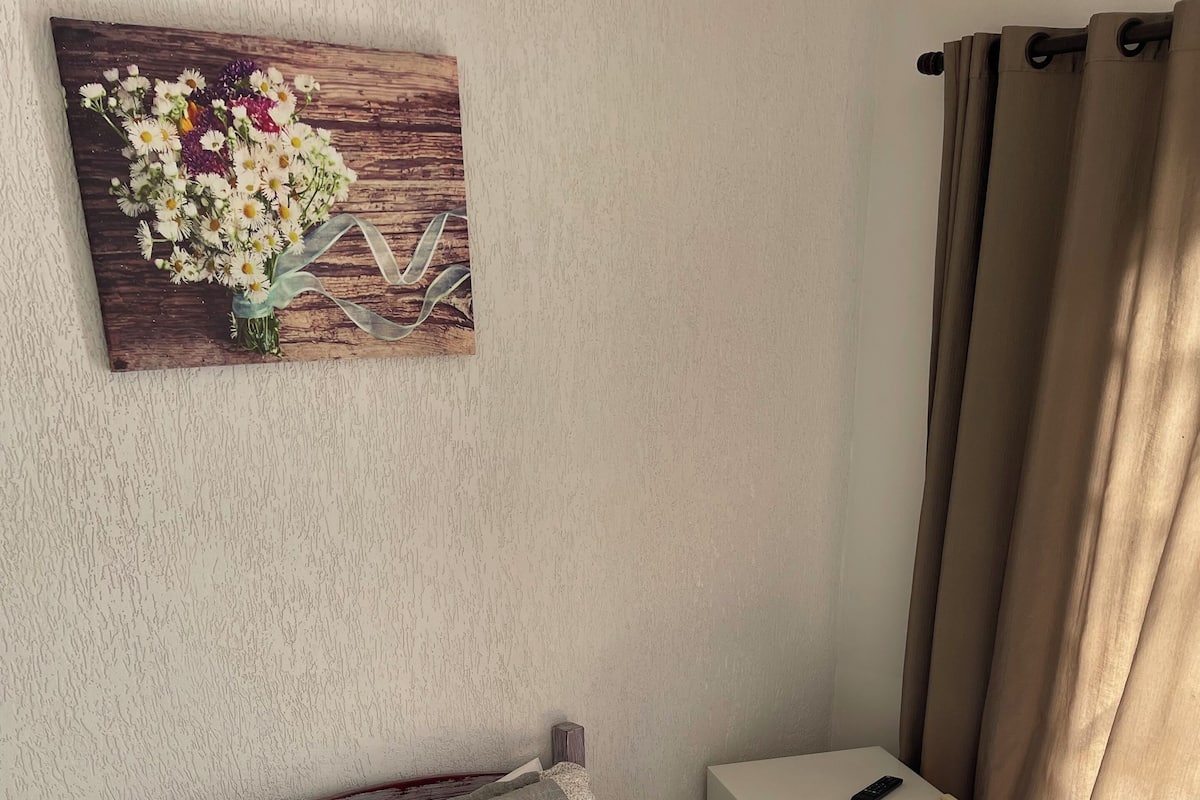 A bedroom with a bed, pillows, a bedside table with a remote control, a small white cabinet, and a floral artwork on the wall.