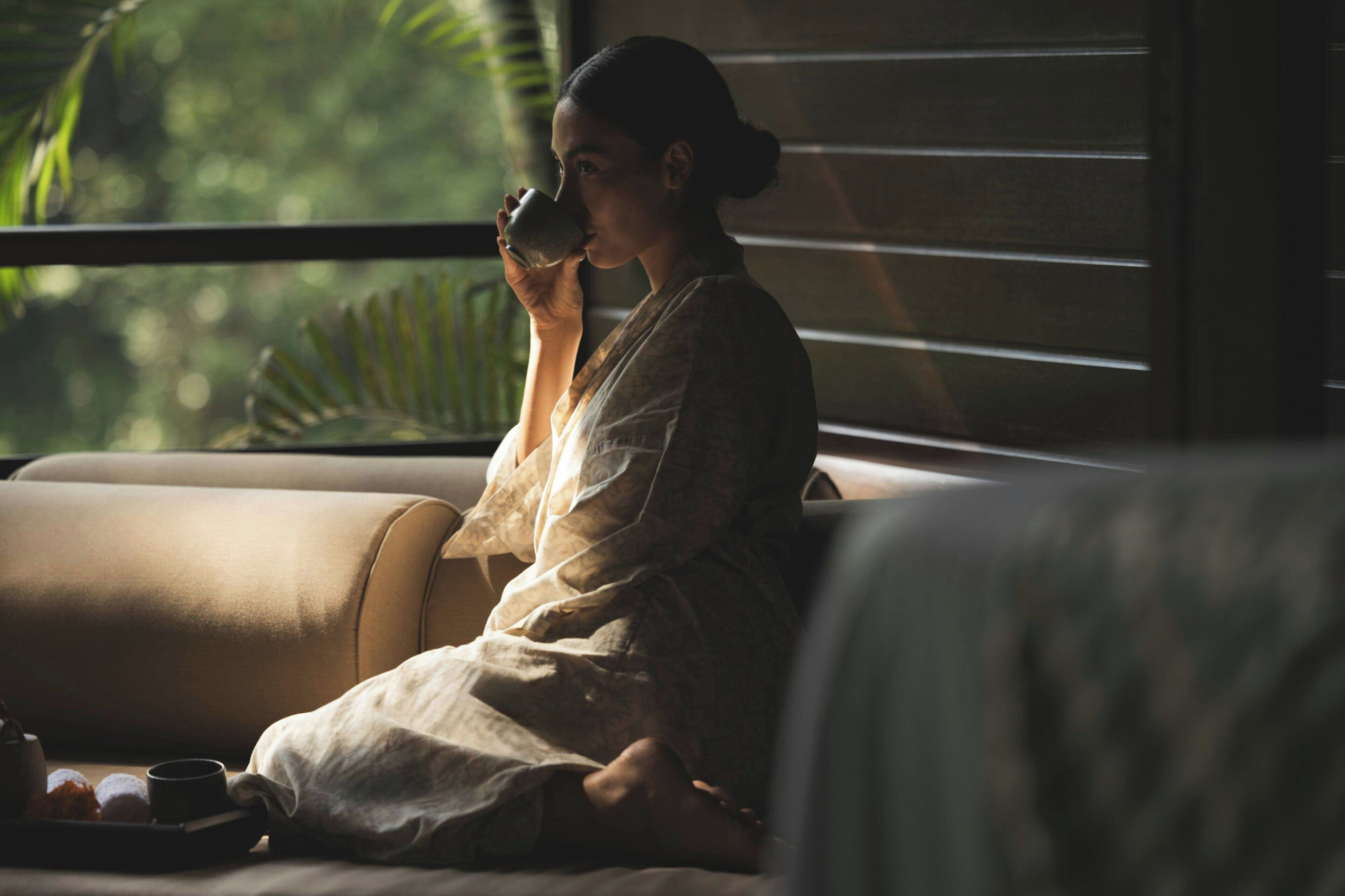 A person sitting on a sofa with a cup, a tray with a teapot and cups, and a window with blinds.