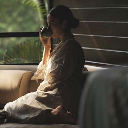 A person sitting on a sofa with a cup, a tray with a teapot and cups, and a window with blinds.