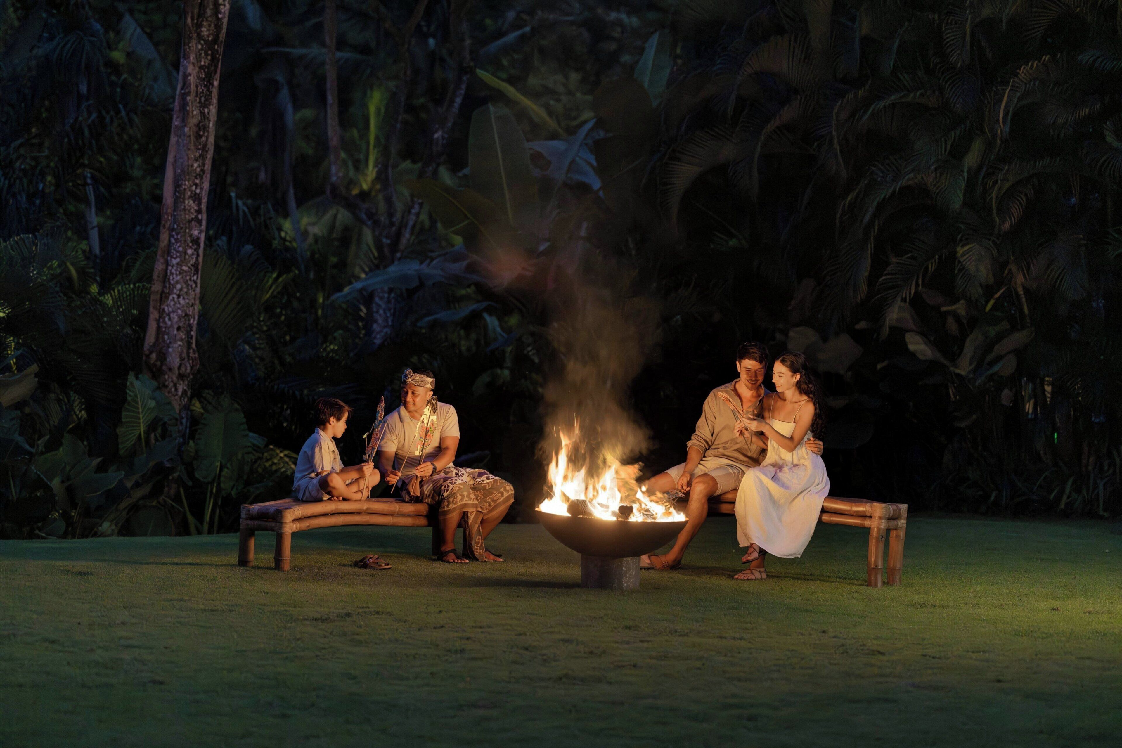 A group of people sitting around a fire pit in a tropical setting with dense foliage in the background.