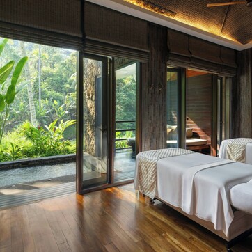 A room with two beds, a large window with a view of greenery, and a wooden floor.
