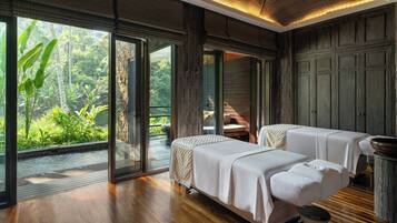 A room with two beds, a large window with a view of greenery, and a wooden floor.