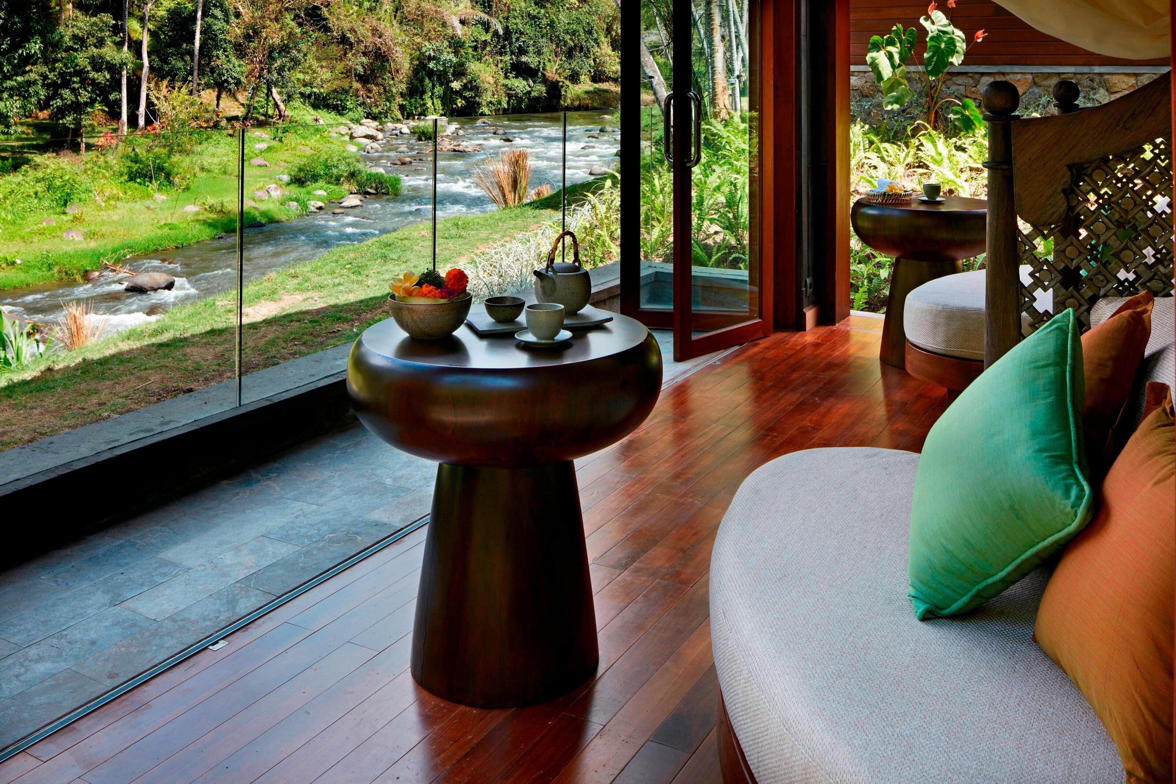 A room with a wooden floor, a sofa with cushions, a table with a bowl of fruit and tea set, and a view of a river through large windows.