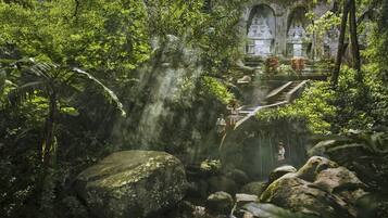 A lush, tropical garden with a waterfall, large rocks, and a stone structure in the background.