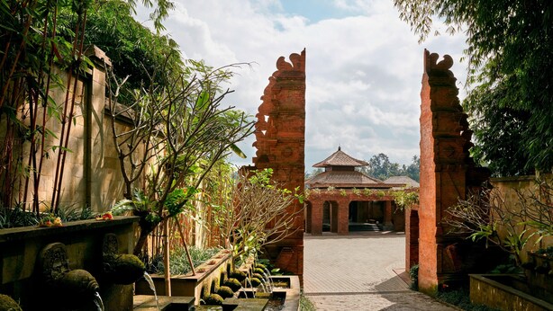 A pathway leading to a traditional building with a tiled roof, surrounded by lush greenery and a water feature with multiple tiers.