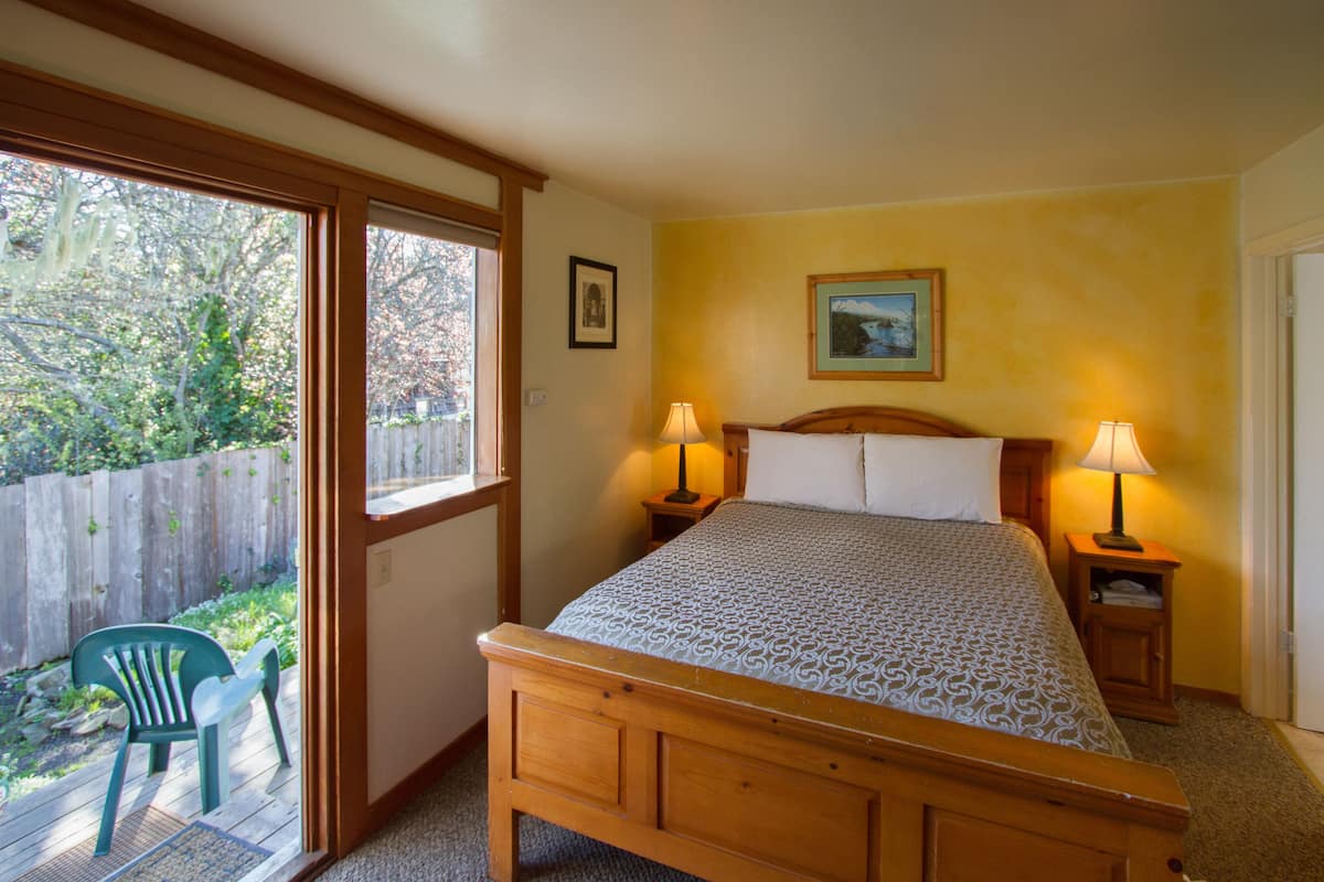 A bedroom with a wooden bed, two bedside tables with lamps, a window with a view of the backyard, and a door to the outside.