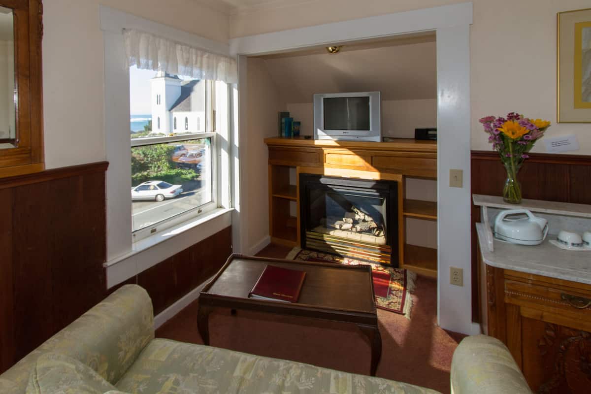 A living room with a fireplace, a television on a stand, a window with a view of a building and a car, and a vase with flowers on a table.