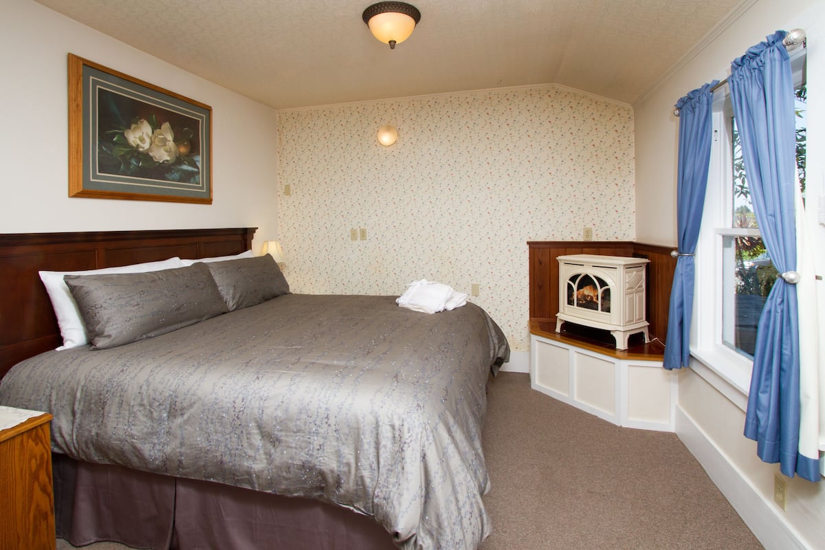 A bedroom with a large bed, a nightstand, a dresser, a wall-mounted lamp, a painting, and a window with curtains.