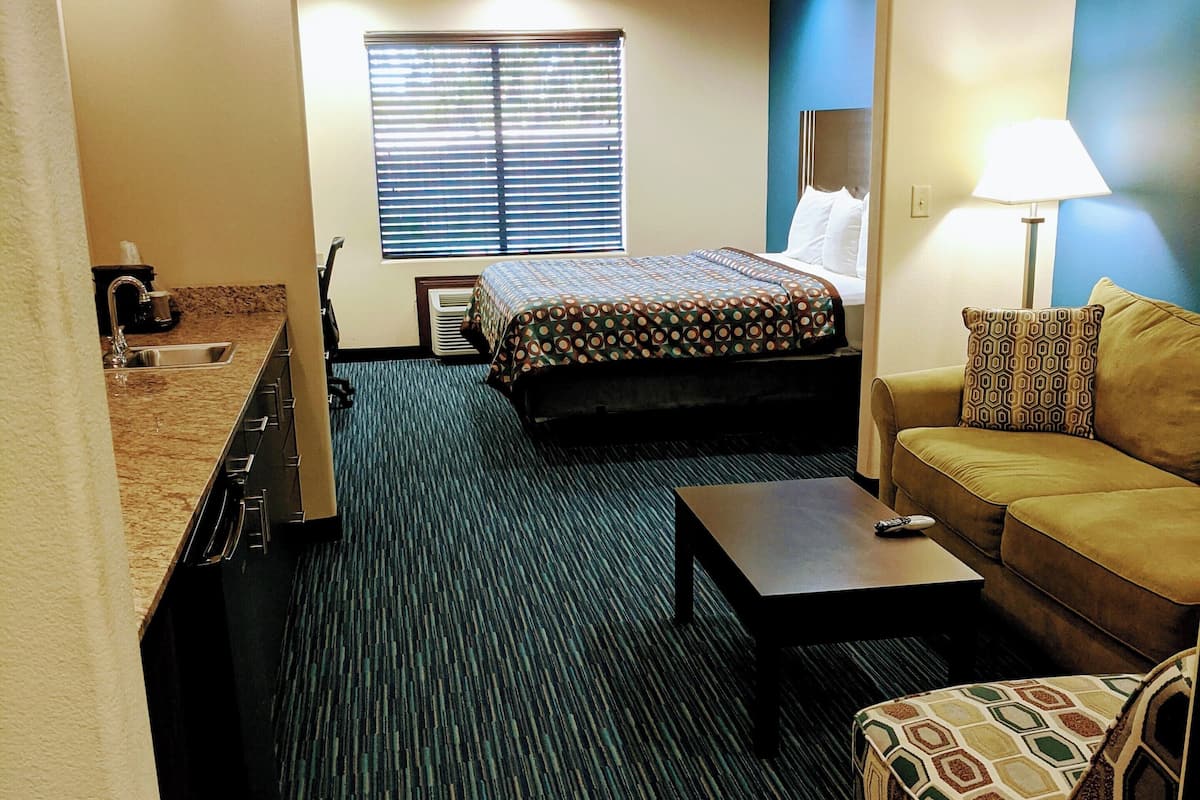 A hotel room with a bed, a sofa, a coffee table, a lamp, and a kitchenette.