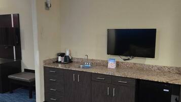 Habitación de hotel compacta con kitchenette, televisor de pantalla plana montado en la pared, cafetera y una pequeña zona de estar con una silla.