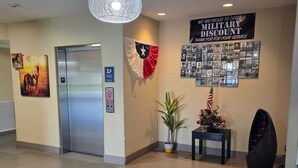 A lobby with a flag, a military discount poster, a wall-mounted photo collage, a potted plant, and a modern sculpture. - Karnes City Lodge  (Karnes City)