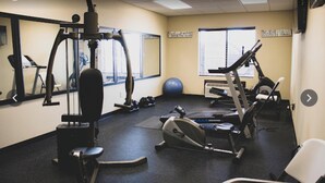 A gym with exercise equipment, a mirrored wall, and a window with blinds. - Karnes City Lodge  (Karnes City)