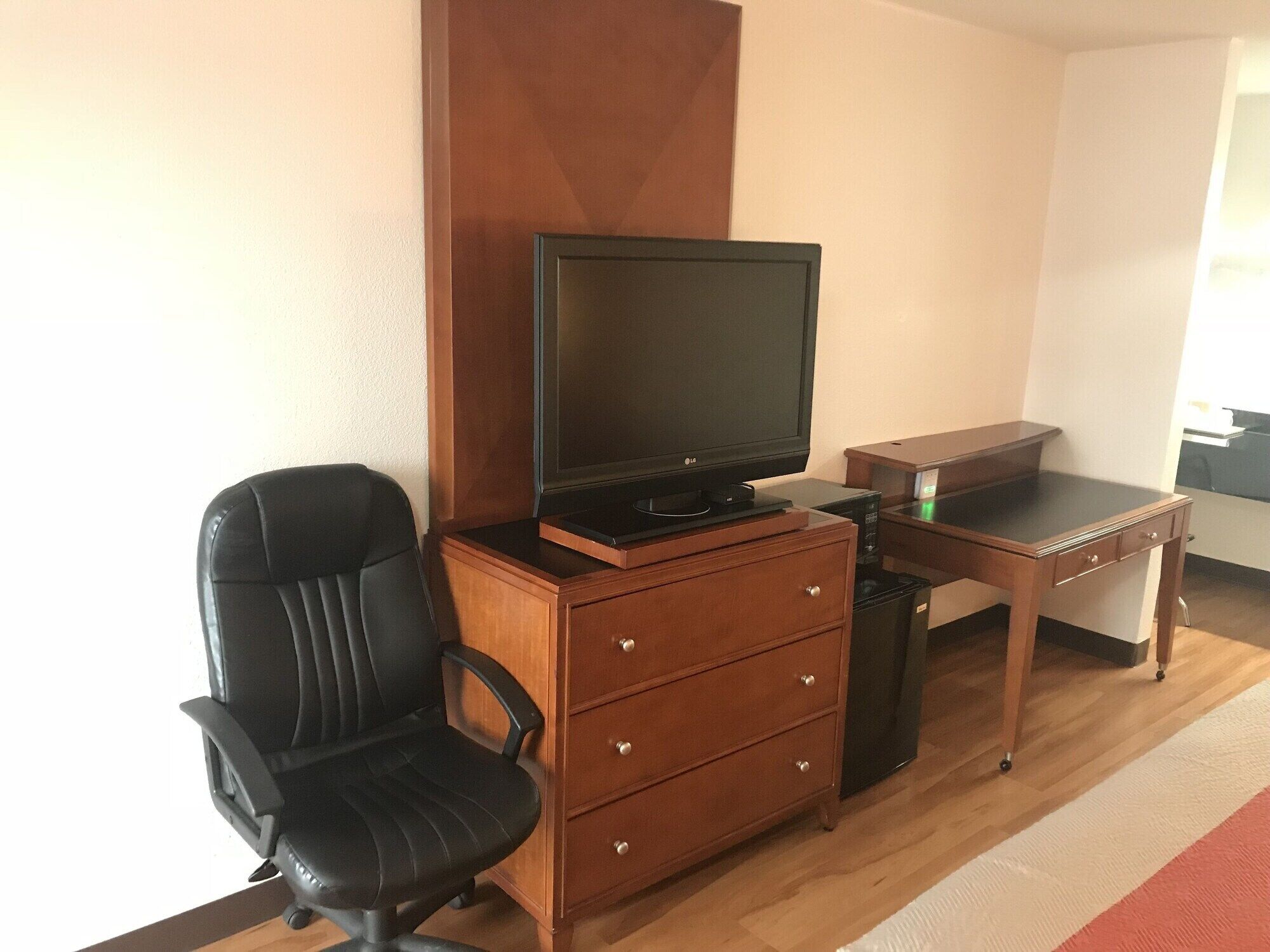 A small room with a wooden desk, a black office chair, a television on a wooden stand, and a small cabinet with drawers.